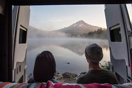 couple sitting at edge of RV at a foggy, placid lake with mountains in background