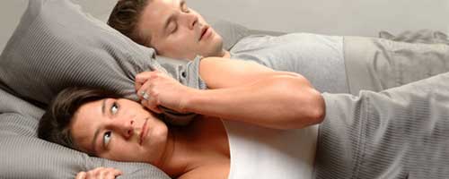woman covering her ears with pillow while laying next to snoring partner