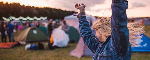 girl with blonde hair and jean jacket dancing hands in the air and tents in background
