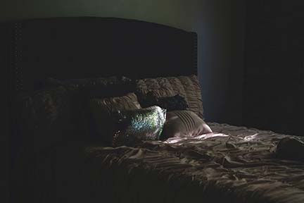 dark bedroom setting with lots of pillows and a sliver of light coming through the blinds