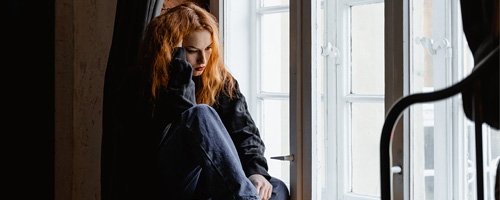 Woman with Seasonal Affective Disorder is sitting on a window sill with an upset look on her face and her hand on her head.